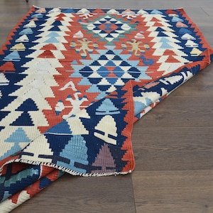 K&ouml;nnte beinhalten: Ein bunter Teppich mit einem geometrischen Muster in Blaut&ouml;nen, Rot, Wei&szlig; und Creme. Der Teppich ist in der Mitte gefaltet, wodurch das komplizierte Design sichtbar wird.
