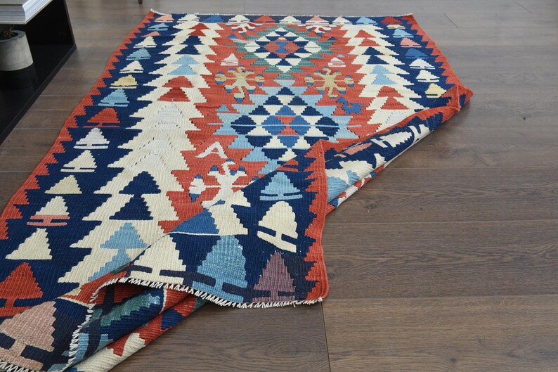 K&ouml;nnte beinhalten: Ein bunter Teppich mit einem geometrischen Muster in Blaut&ouml;nen, Rot, Wei&szlig; und Creme. Der Teppich ist in der Mitte gefaltet, wodurch das komplizierte Design sichtbar wird.