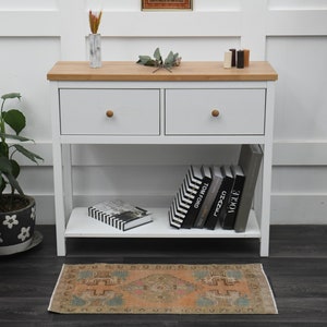 Puede incluir: Mesa consola blanca con dos cajones y un estante inferior. La mesa tiene una tapa de madera y está decorada con libros, una planta y un jarrón de flores.