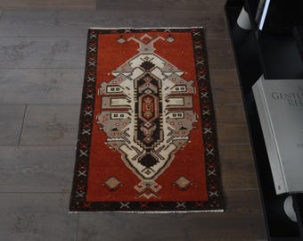May include: A rectangular, hand-woven rug with a central geometric design in shades of brown, beige, and gray, set against a rust-colored background. The rug has a dark brown border with repeating cross-like patterns. The rug is approximately 36 inches long and 24 inches wide.