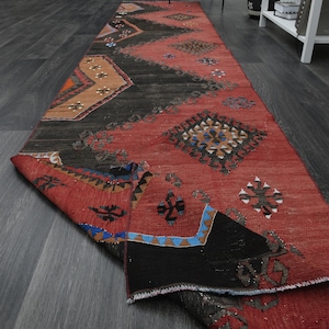 May include: A red, black, and blue patterned runner rug with a geometric design. The rug is folded in half and is lying on a dark gray wood floor.