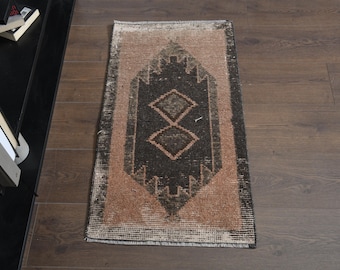 Könnte beinhalten: Ein rechteckiger Vintage-Teppich mit einem zentralen geometrischen Muster in Schwarz und Braun, umrahmt von einem hellbraunen Rand. Das abgenutzte Aussehen des Teppichs deutet auf Alter hin und trägt zu seinem einzigartigen Charakter bei. Der Teppich liegt auf einem Holzboden.