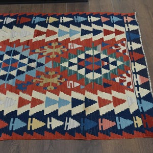 K&ouml;nnte beinhalten: Ein bunter Teppich mit einem geometrischen Muster in Rot-, Blau-, Wei&szlig;- und Gr&uuml;nt&ouml;nen. Der Teppich hat ein traditionelles Design mit einer zentralen Diamantform und sich wiederholenden Dreiecken.