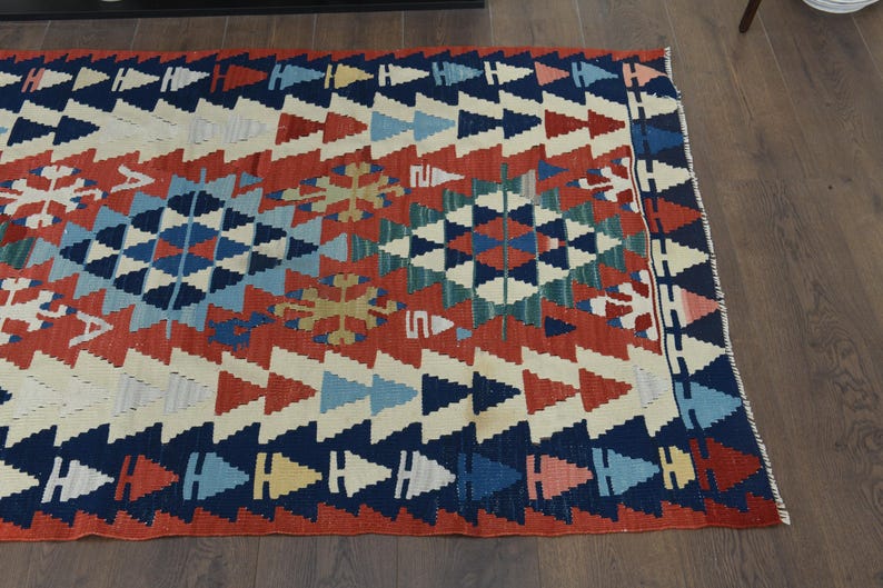 K&ouml;nnte beinhalten: Ein bunter Teppich mit einem geometrischen Muster in Rot-, Blau-, Wei&szlig;- und Gr&uuml;nt&ouml;nen. Der Teppich hat ein traditionelles Design mit einer zentralen Diamantform und sich wiederholenden Dreiecken.