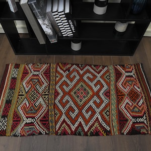 May include: A rectangular, hand-woven rug with a geometric pattern. The rug features a central diamond design in shades of red, orange, and white, framed by intricate borders in various colors. The rug is placed on a wooden floor.