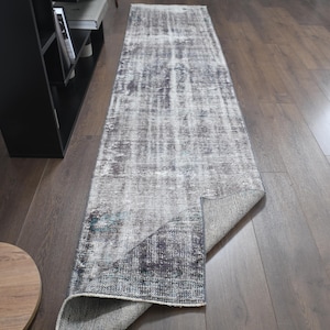 May include: A rectangular, distressed-look rug with a faded gray and blue pattern. The rug is partially rolled up, revealing a lighter gray underside. It is placed on a dark wood floor.
