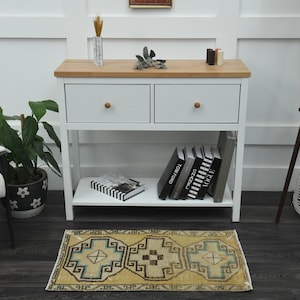 May include: A white console table with two drawers and a lower shelf. The table has a wood top and is decorated with a small plant, books, and a rug. The rug is beige with a blue and brown geometric pattern.