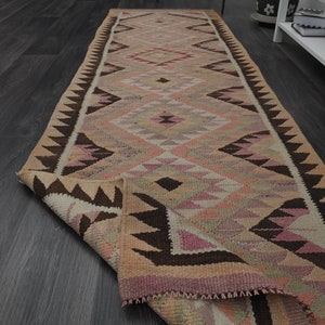 May include: A long, rectangular rug with a geometric pattern in shades of beige, pink, and brown. The rug features diamond and triangle shapes, with a dark brown border. The rug is partially rolled up, revealing the underside.