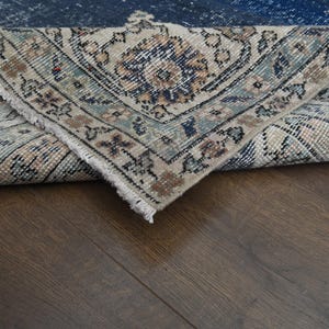 May include: A close-up of a folded rug with a blue and cream patterned design. The rug is folded over on itself, revealing the underside of the rug.