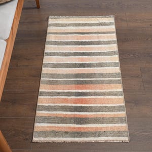 May include: A striped rug in peach, grey, and white, with a fringed edge. The rug is rectangular and lies on a dark wooden floor. The design features horizontal stripes, suitable for various interior settings. The rug's dimensions are in centimeters.