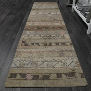 May include: A long, rectangular rug with a faded, vintage look. The rug has a beige background with brown, pink, and blue geometric patterns. The rug is made of woven fabric and has a frayed edge.