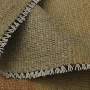 May include: Close-up of a woven rug with a fringe border. The rug is a light brown color with a subtle pattern.