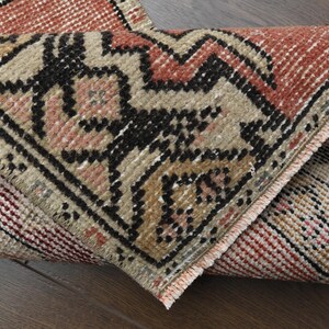 May include: Close-up of a folded, vintage rug with a geometric pattern. The rug features a combination of colors, including red, black, beige, and pink. The design includes zigzags and floral elements, with a fringed edge.
