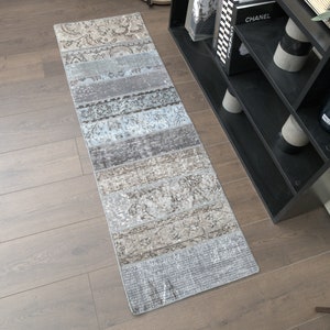 May include: A long, rectangular runner rug with a faded, vintage look. The rug has a light blue, gray, and brown color scheme with a patterned design. The rug is laid out on a wooden floor.