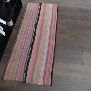 May include: A long, narrow, striped rug with pink, brown, and purple stripes. The rug is made of woven fabric and has a fringed edge.