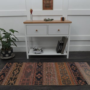 May include: A white console table with two drawers and a lower shelf. The table has a wood top and is decorated with a small potted plant, books, and a rug with a colourful geometric pattern.