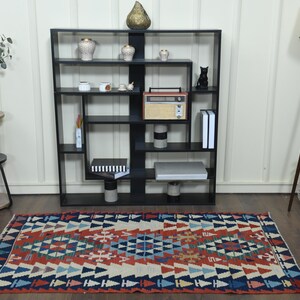 K&ouml;nnte beinhalten: Ein bunter Teppich mit geometrischem Muster in Rot-, Blau- und Wei&szlig;t&ouml;nen. Der Teppich liegt auf einem Holzboden vor einem schwarzen B&uuml;cherregal mit verschiedenen Dekorationsartikeln.