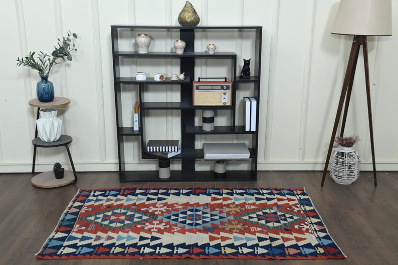 K&ouml;nnte beinhalten: Ein bunter Teppich mit geometrischem Muster in Rot-, Blau- und Wei&szlig;t&ouml;nen. Der Teppich liegt auf einem Holzboden vor einem schwarzen B&uuml;cherregal mit verschiedenen Dekorationsartikeln.