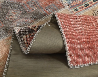 May include: A section of a vintage patchwork rug with a distressed appearance. The rug features a mix of colors, including red, brown, beige, and gray, with visible stitching along the edges. The rug is folded over, revealing a brown backing.