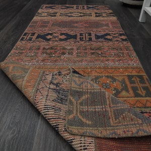 May include: A colourful, hand-woven rug with intricate geometric patterns in shades of red, orange, blue, and black. The rug is folded in half, revealing the back side, which is a lighter shade of brown.