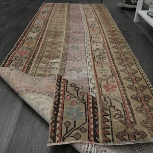 Può includere: Un tappeto da runner vintage con sfondo beige e motivi intricati in tonalità di marrone, rosso e rosa. Il tappeto ha un bordo con frange ed è piegato a metà, rivelando il lato inferiore.