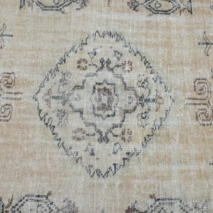 May include: A woven rug featuring a light tan background with a central diamond-shaped medallion and repeating geometric floral motifs in dark grey, brown, and muted black tones across the entire textile surface.