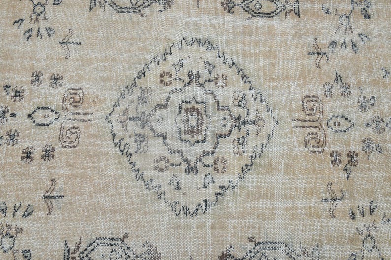 May include: A woven rug featuring a light tan background with a central diamond-shaped medallion and repeating geometric floral motifs in dark grey, brown, and muted black tones across the entire textile surface.