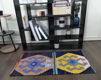 May include: A black and blue patterned rug with yellow, red, and white accents. The rug is on a wooden floor in front of a black bookshelf with various items on the shelves.