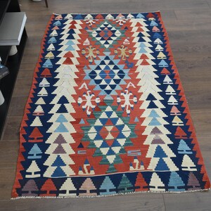 K&ouml;nnte beinhalten: Ein bunter Teppich mit einem geometrischen Muster in Rot-, Blau-, Wei&szlig;- und Gr&uuml;nt&ouml;nen. Der Teppich hat eine zentrale Diamantform mit sich wiederholenden Dreiecken und floralen Motiven.