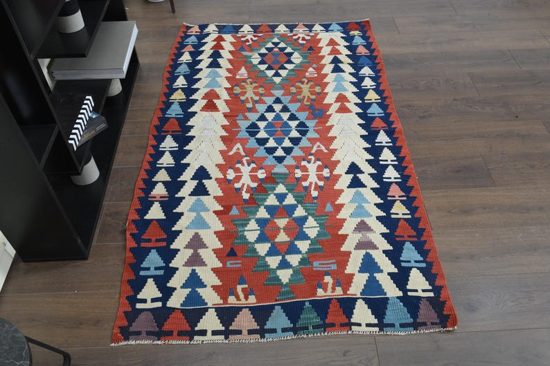 K&ouml;nnte beinhalten: Ein bunter Teppich mit einem geometrischen Muster in Rot-, Blau-, Wei&szlig;- und Gr&uuml;nt&ouml;nen. Der Teppich hat eine zentrale Diamantform mit sich wiederholenden Dreiecken und floralen Motiven.