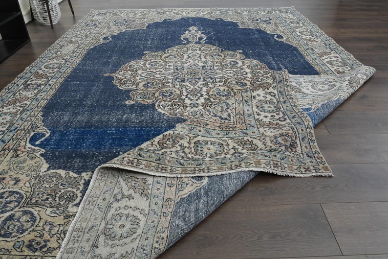 May include: A vintage rug with a blue center and a cream-colored border. The rug has a floral pattern and is folded in half.