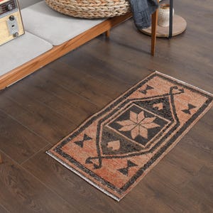 May include: A small, rectangular rug with a geometric pattern in shades of orange, black, and beige. The rug features a central starburst design and is placed on a dark wooden floor. The rug is likely handmade and adds a touch of warmth to the room.