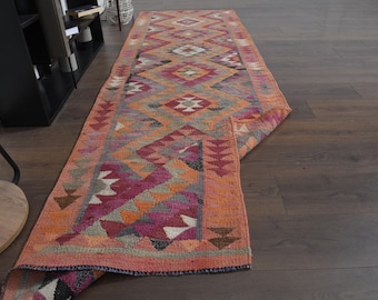 May include: A rectangular, hand-woven runner rug with a geometric pattern. The rug features a vibrant color palette of orange, pink, red, and gray. The design includes diamond and triangle shapes. The rug is displayed on a dark wood floor.