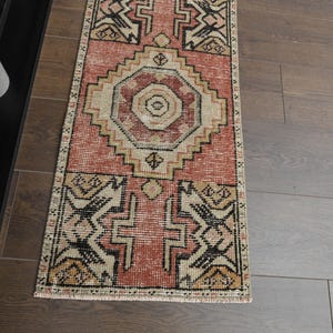 Könnte beinhalten: Ein rechteckiger Teppich im Vintage-Stil mit einem zentralen Medaillon-Design in Pfirsich-, Beige- und Schwarztönen. Der Teppich weist geometrische Muster und einen Used-Look auf, geeignet für einen Flur oder Eingangsbereich. Der Teppich liegt auf einem Holzboden.