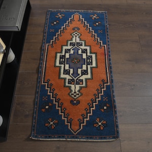 Könnte beinhalten: Ein rechteckiger Teppich mit einem leuchtend orangefarbenen zentralen Rautenmotiv auf tiefblauem Grund. Das Design umfasst geometrische Muster in Weiß, Dunkelblau und Orange sowie kleine blumenartige Akzente in den Ecken.