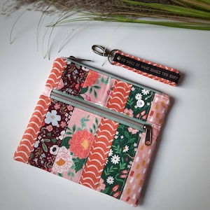 Könnte beinhalten: Eine gepolsterte Tasche aus Patchwork-Stoff mit floralem und geometrischem Muster. Die Tasche hat einen Reißverschluss und einen abnehmbaren Schlüsselanhänger mit dem Text "Tu mehr von dem, was dich glücklich macht".