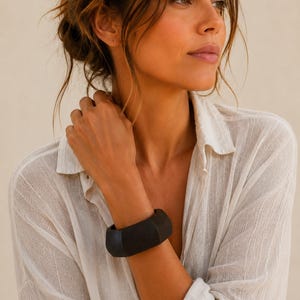May include: A wide, matte black bangle bracelet with a faceted, geometric design. The bracelet features a solid, dark construction with distinct angular edges that create a structured look around the wrist, contrasting with the light-colored fabric.