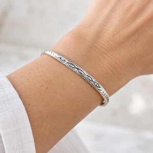 May include: A slim silver cuff bracelet featuring an oxidized finish and a repeating geometric pattern of small, etched diamond shapes along the center, bordered by delicate dotted lines that run the length of the polished metal band.