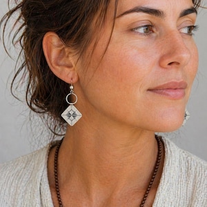 Peut inclure: Boucles d'oreilles en argent avec un motif géométrique. Chaque boucle d'oreille présente un pendentif en forme de losange avec un motif noir et blanc, suspendu à un petit anneau en argent. La personne porte un pull de couleur claire.