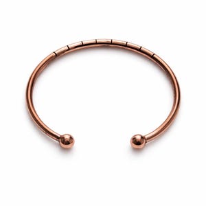 Op de afbeelding: Een koperkleurige manchetarmband met een minimalistisch design. De open armband heeft een gladde, afgeronde band met kleine, decoratieve lijnen die aan de bovenkant zijn geëtst. Elk uiteinde van de armband heeft een klein, bolvormig detail.