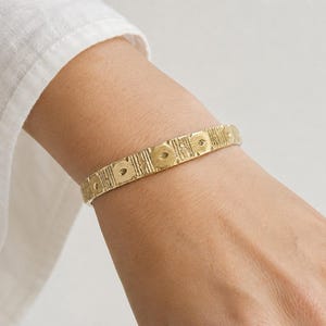 May include: A gold-toned metal bangle bracelet featuring a repeating pattern of circular indentations and vertical textured lines along the entire outer surface. The bracelet has a consistent width and a matte finish, encircling the wrist area.