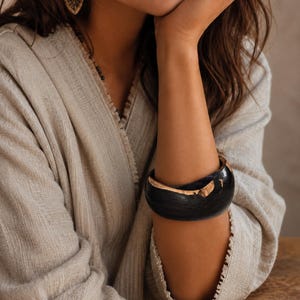 Peut inclure: Un large bracelet jonc incurvé fabriqué en bois sombre poli, présentant des accents de grain de bois naturel clair le long des bords. Le bracelet possède une surface lisse et arrondie ainsi qu'une construction épaisse et solide.