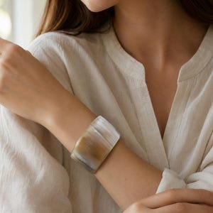 Op de afbeelding: Een brede armband met een glad, gepolijst oppervlak en een natuurlijk verloop van doorschijnende witte, grijze en warme amberkleurige tinten. De armband heeft een gebogen, solide structuur die om de pols sluit en organische kleurvariaties toont.