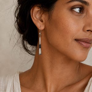 Op de afbeelding: Een zilveren hangende oorbel met een slank, taps toelopend cilindrisch ontwerp en een gepolijste metalen afwerking. De oorbel is voorzien van horizontale zwarte banden rond het onderste gedeelte en hangt aan een eenvoudige zilveren haak.