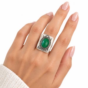May include: A rectangular silver-toned ring featuring a central oval-shaped green stone. The metal setting has an oxidized finish with intricate etched detailing surrounding the stone, creating a textured appearance on the face of the ring.