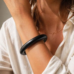 May include: A black, smooth, round bangle bracelet. The bracelet is worn on the arm of a person wearing a white shirt and a delicate gold necklace. The bracelet is the focal point of the image.