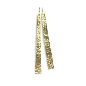 May include: A pair of long, hammered gold earrings. The earrings have a textured surface and a rectangular shape. The earrings hang from silver hooks and are displayed against a white background.