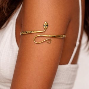 Op de afbeelding: Goudkleurige armmanchet in een slangenontwerp. De manchet wikkelt zich om de bovenarm en heeft een slangenkop met zwarte accenten en een gebogen staart. De sieraden worden om de arm van een persoon gedragen.