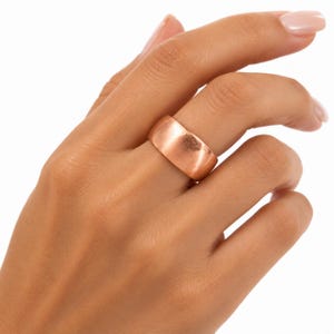 May include: A wide, polished copper band ring with a smooth, rounded surface and a warm, metallic reddish-brown finish. The ring features a simple, solid construction with a consistent width throughout, showcasing a reflective, lustrous appearance.