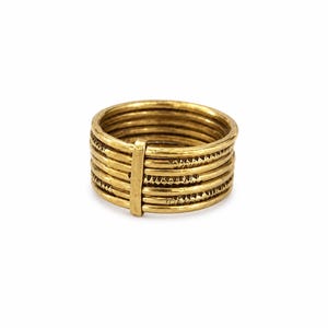 Può includere: Un anello multi-fascia color oro composto da sette sottili anelli sovrapposti, tenuti insieme da una singola barra metallica verticale. La superficie degli anelli presenta un motivo triangolare testurizzato lungo la circonferenza.